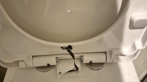 The lifted lid of a white toilet seat, with a black camera tucked under its rim and a wire leading to another part of it wrapped in kitchen roll