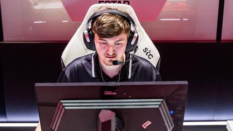 A young man wearing a look of intense concentrations sits in front of a black computer monitor. He's wearing a large, expensive-looking gaming headset with a microphone attachment, and sitting in a large, white gaming chair.