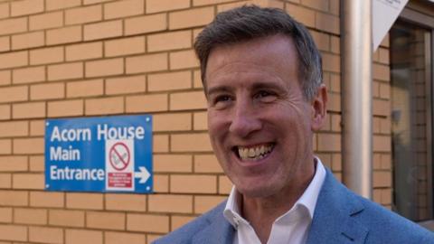 Anton Du Beke is wearing a blue suit jacket over a white shirt. He is smiling in front of a cream brick building that has a blue sign saying "Acorn House main entrance" on the wall.