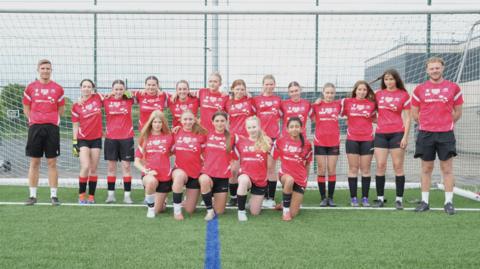 A group of girls in red T-shirts and black shorts in a line, with five girls crouched in front. A male coach is either side of them, with a football goal behind.