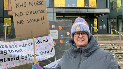 Beth Crisan, a mother of a child who attends Steps Ahead Nursery, wearing a grey padded raincoat and a colourful knitted beanie hat. She is standing near the steps outside County Hall, holding a cardboard sign which says "full-time NHS nurse, no childcare, no work".