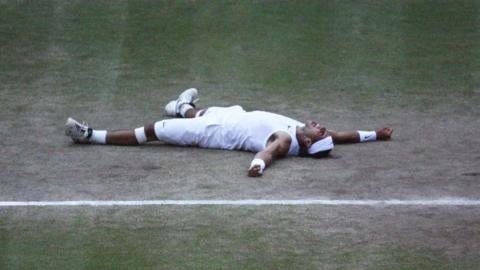 A man lays down on a grass tennis court in tennis whites, with his arms and legs wide in celebration.
