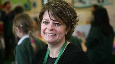 Ruth Perry smiling and wearing a black top with green lanyard. She is in a classroom with blurred children in the background.