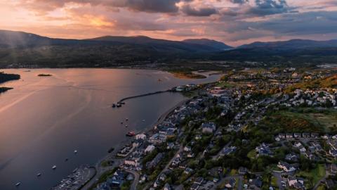 An aerial shot showing multiple buildings on a green grassy area, next to a seafront. Hills fill the background, with a sunset setting in. 
