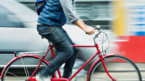 A man on a red bicycle is pictured travelling at pace near a moving car. He is wearing a blue jacket and has navy jeans. He has a black backpack on.