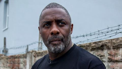 Idris Elba looks away from camera with a barbed fence atop a wall in the backround