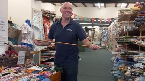 Bryan Machin wearing a blue polo shirt and standing in the haberdashery store. He is holding a metre stick