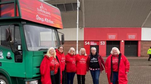 A group of six volunteers from the Sunderland Community Soup Kitchen stand in front of a green lorry which has the name of the charity on a red/orange sign above the cab. They are outside the Stadium of Light, with part of a stand and closed turnstiles behind them.