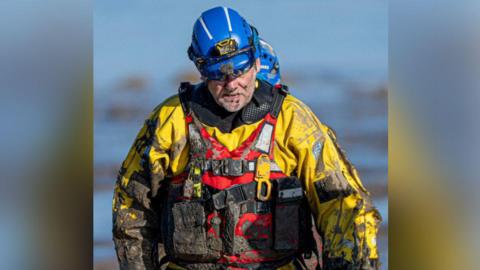 A coastguard crew in specialist uniform covered in mud.