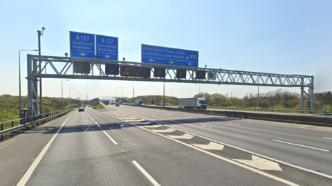 Part of the M25 motorway which has four lanes, between junction 29 and junction 30 in Essex. The sky is blue and blue overhead signs are directing traffic to Southend and Basildon and onwards to Tilbury and the Dartford Crossing.