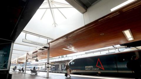 A train platform with a green Avanti West Coast train pulling into the station. The platform is bright and lit up by long rectangular skylights that are inset in the wooden ceiling panels. There are people on the platform.