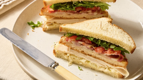 The image shows a neatly prepared sandwich cut into triangular halves, stacked slightly apart on a light-colored ceramic plate. The sandwich is made with soft white bread, with a golden-brown crust and a fluffy interior. Inside the sandwich, there are visible layers of sliced deli meat, fresh green leafy vegetables and slices of red tomato, secured with a cocktail stick