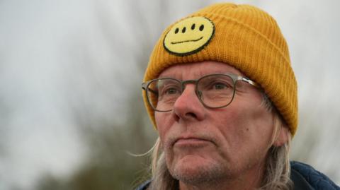 Neil has shoulder length blonde hair and is wearing a dark yellow woolen hat and dark rimmed glasses.