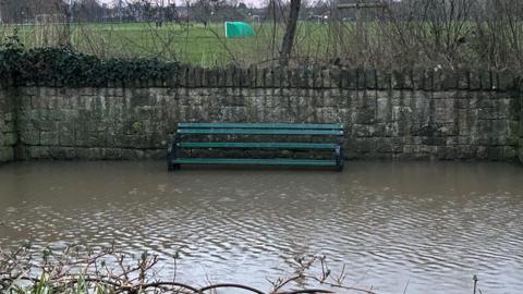 A picture of a green bench which has water up to the seated area. In front of it the stream has flooded. In the background is a football ground which has a green goal. 
