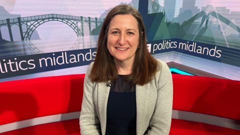 Julia Buckley has brown hair just past her shoulders and is wearing a black top with a grey suit jacket. She is sitting on a red sofa and in the background there is a banner that reads "politics midlands"