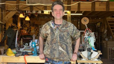 A man wearing a multi-coloured T-shirt and jacket is standing in front of a wooden workbench. A range of tools and equipment for shoemaking can be seen behind him.