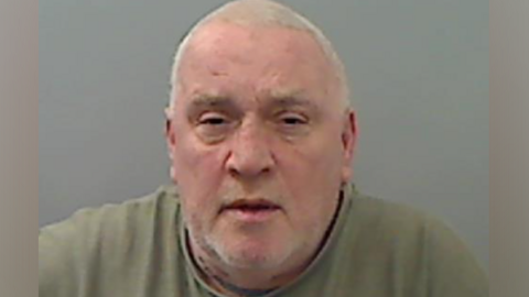 Edwin Youll with short white hair, wearing a green top, looks directly at the camera in a police mugshot photo, with a grey background.