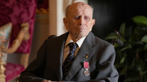 Norman is wearing a suit and a medal is pined on. He's sitting down.