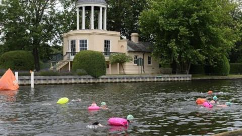 Swimmers with bright pink, yellow and orange floats in a river with pale green hats on a cream building can be seen on the edge of the river.