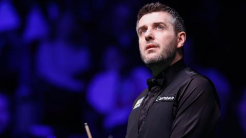 Mark Selby looks upwards during a snooker match, wearing a black shirt with sponsor logos