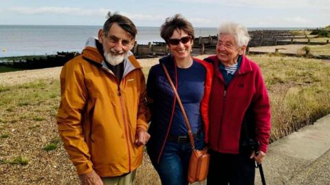 Colin, Christine and Beryl Bell are standing in front of a beach. Colin - an older, bearded man on the left of the three is smiling. He's wearing an orange raincoat. Christine, in the middle of the three, is wearing a blue coat and sunglasses. She has short brown hair. On the right, Beryl is an older woman in a red fleece coat. She is smiling at Christine and holding onto a walking stick.