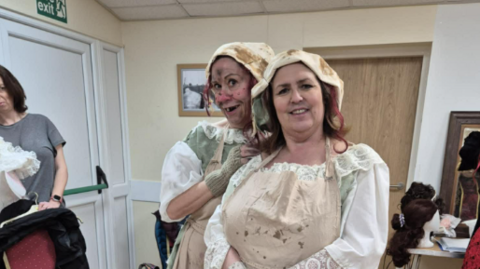 Two women in dirty white dresses in a room with beige walls and white doors