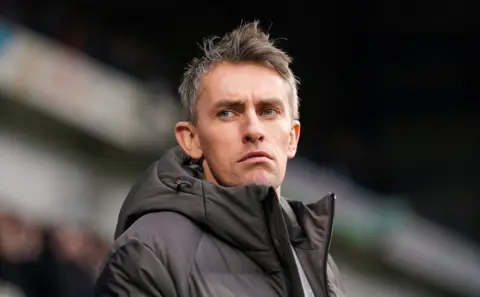 Close-up shot of Kieran McKenna, manager of Ipswich Town, wearing a dark grey hooded puffer jacket and looking off-camera with a focused expression during a match.