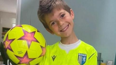 Alfie Tollett pictured wearing a bright yellow Gillingham Football Club jersey and holding a yellow football with purple stars all around it. He has brown hair and is smiling at the camera. He is in a house with light blue walls.