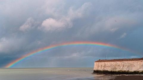 A shallow rainbow in a dark sky above the sea and a sea cliff