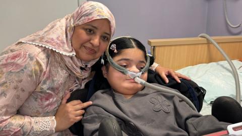 Maham looks at her iPad as she sits with her mother, Nomia. She is wearing a device which helps with ventilation which happens twice a day and through the night.
