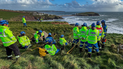 A group of coastguard rescue officers wearing high-visibility clothing and safety helmets take part in a cliff rescue training exercise on a coastal headland overlooking the sea. Ropes, rescue equipment and anchor systems can be seen on the grass as volunteers practise techniques above a rugged shoreline and surf-filled bay.