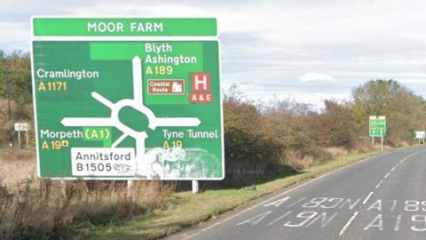 The A189 leading up to Moor Farm roundabout. A large green sign reads Moor Farm and shows the layout of the approaching roundabout. It is a dual carriageway.