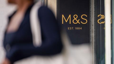 The M&S logo is seen pictured next to a note saying 'est. 1884' on the side of a Marks and Spencer store with an out-of-focus anonymous shopper holding a canvas bag in the foreground, in London on 1 May