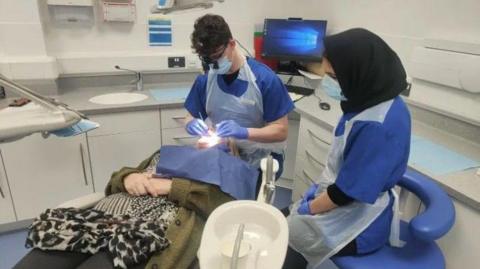 A patient is seated in a dentist's chair. A dental student wearing scrubs and PPE is looking in her mouth. Another student, also wearing scrubs and PPE, is looking on