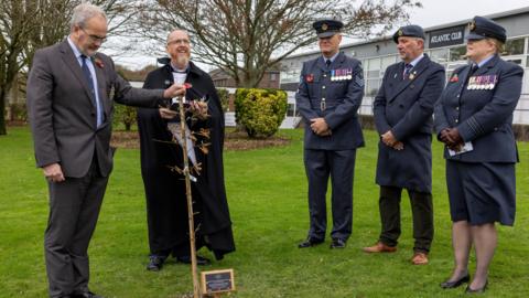 A small ceremonial gathering outdoors on the grass. Five people stand around the newly planted tree, which has a commemorative plaque at its base.