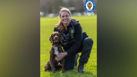 A woman in police uniform sits on the grass with a Chocolate Sprocker dog in front of her.