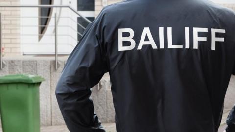 Rear view of a male bailiff standing with his hands on his hips at the entrance of a house.