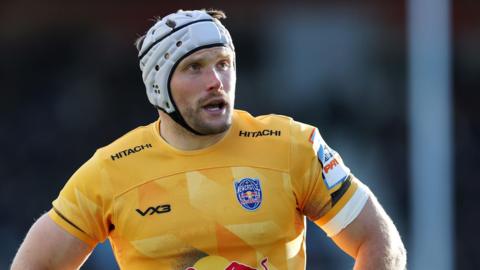 A close up shot of Ollie Leatherbarrow looking onward mid game for Newcastle Red Bull. He wears a white scrum cap and a yellow playing shirt. 