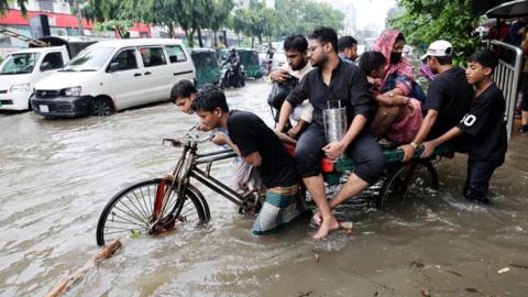Several people push a heavily laded tricycle through floodwaters in Dhaka