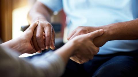 Carer holding the hands of a patient who is sitting in a chair. No faces can be seen, with the picture focused on the hands.