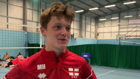 A man wearing a red zip coat stood on a volleyball court in a hall