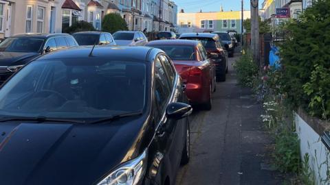 A row of cars parked on a pavement on a narrow street