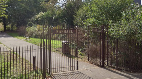Metal gates at the entrance to Raphael Park in Romford, with a paved path leading into the park and trees and greenery beyond.