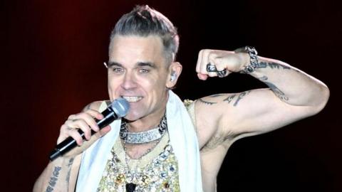 Robbie Williams sings into a microphone on stage. He smiles wearing a sparkly vest