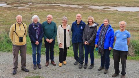 Four men and four women look into the camera and smile while standing on the site of the Rye Harbour Nature Reserve.