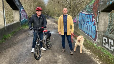 A man wearing a black coat and red cycling helmet sits on a bike. To his left shoulder a man wearing a beige jacket stands with a light brown coloured dog. They are stood on a footbridge with graffiti on either side.