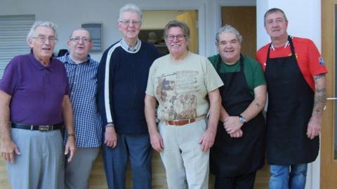 Six men are standing together and smiling next to the charity's kitchen. Two of them are wearing dark-coloured aprons. 