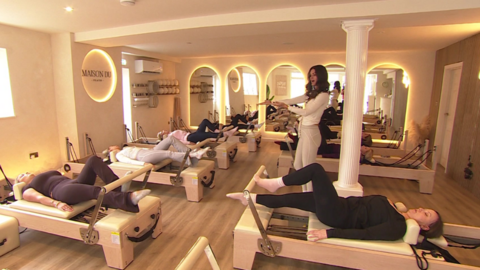 A Reformer Pilates studio with seven machines, each one being used by a woman, and a female instructor stood in the middle of the room