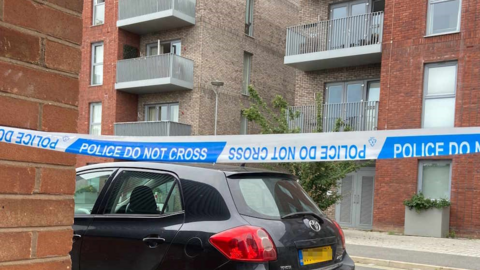 A black car parked by a set of flats. There is police tape in front of the car and flats and it reads Police do not cross.