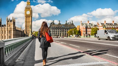 Rear-view of a woman walking along Westminster Bridge. She is walking in bright sunshine towards the Houses of Parliament and Portcullis House, has long brown hair and is dressed in black leggings, a black jacket and brown flat ankle boots and is carrying a red tote bag.
